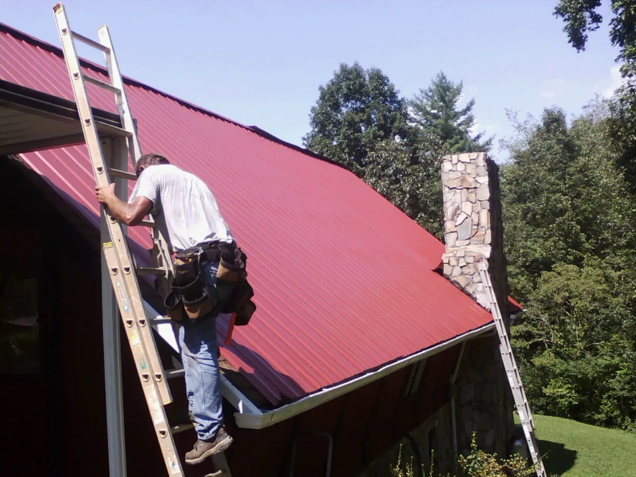 Roofer on a ladder beside a red metal roof and stone chimney — Soffit & Fascia Repair by Roofing Brothers in Sedley